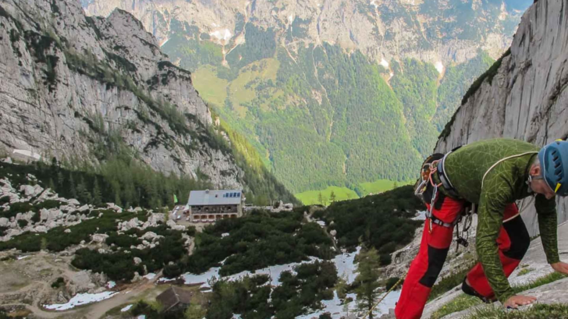 Kletterer steigt eine geneigte Kalkfelsplatte auf, im Hintergrund eine Berghütte mit Satteldach in den Berchtesgadener Alpen.