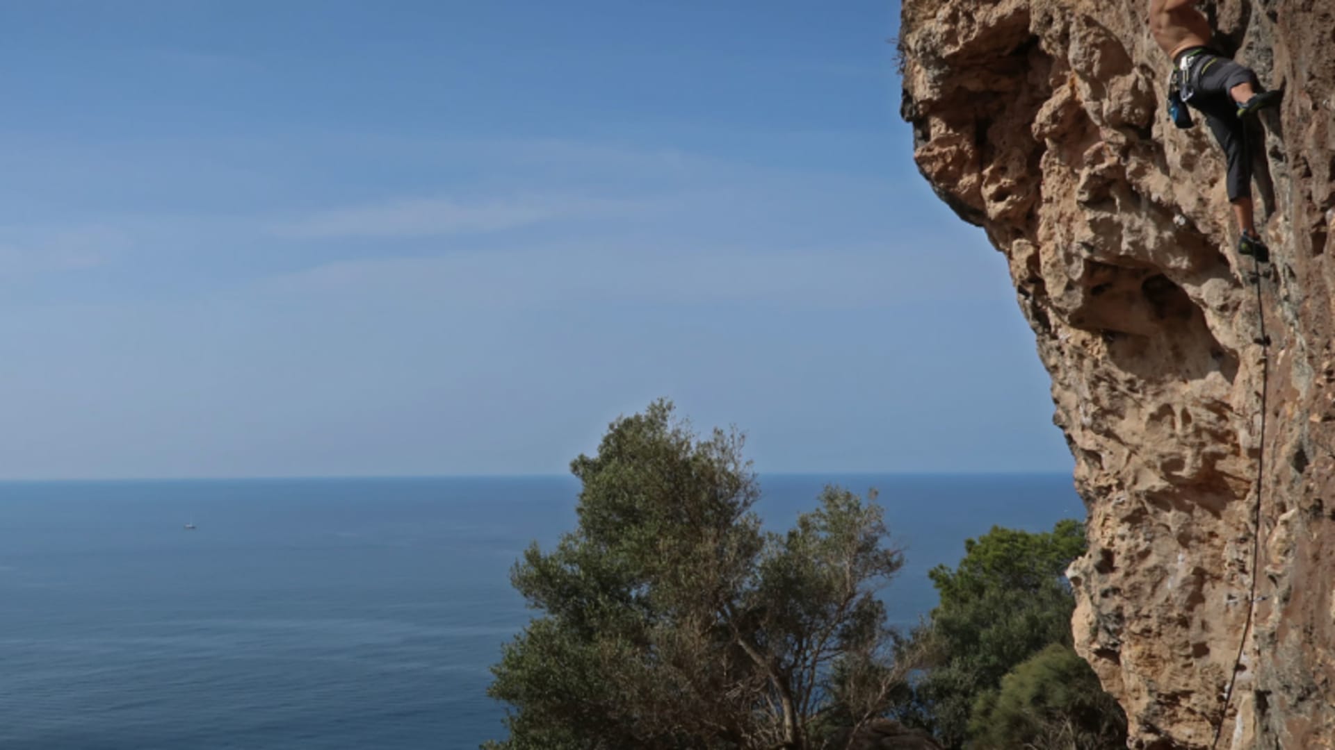 Kletterer steigt senkrechte Kalksteinwand auf Mallorca hoch, im Hintergrund eine Meeresbucht mit steil abfallenden Klippen.