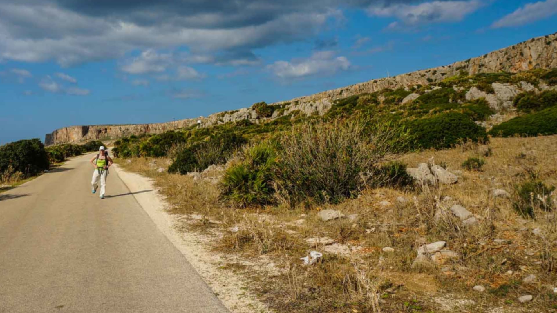 Wanderer auf asphaltierter Küstenstraße in Sizilien, flankiert von Oleander, Kakteen und Kalksteinfelsen im Sommer.