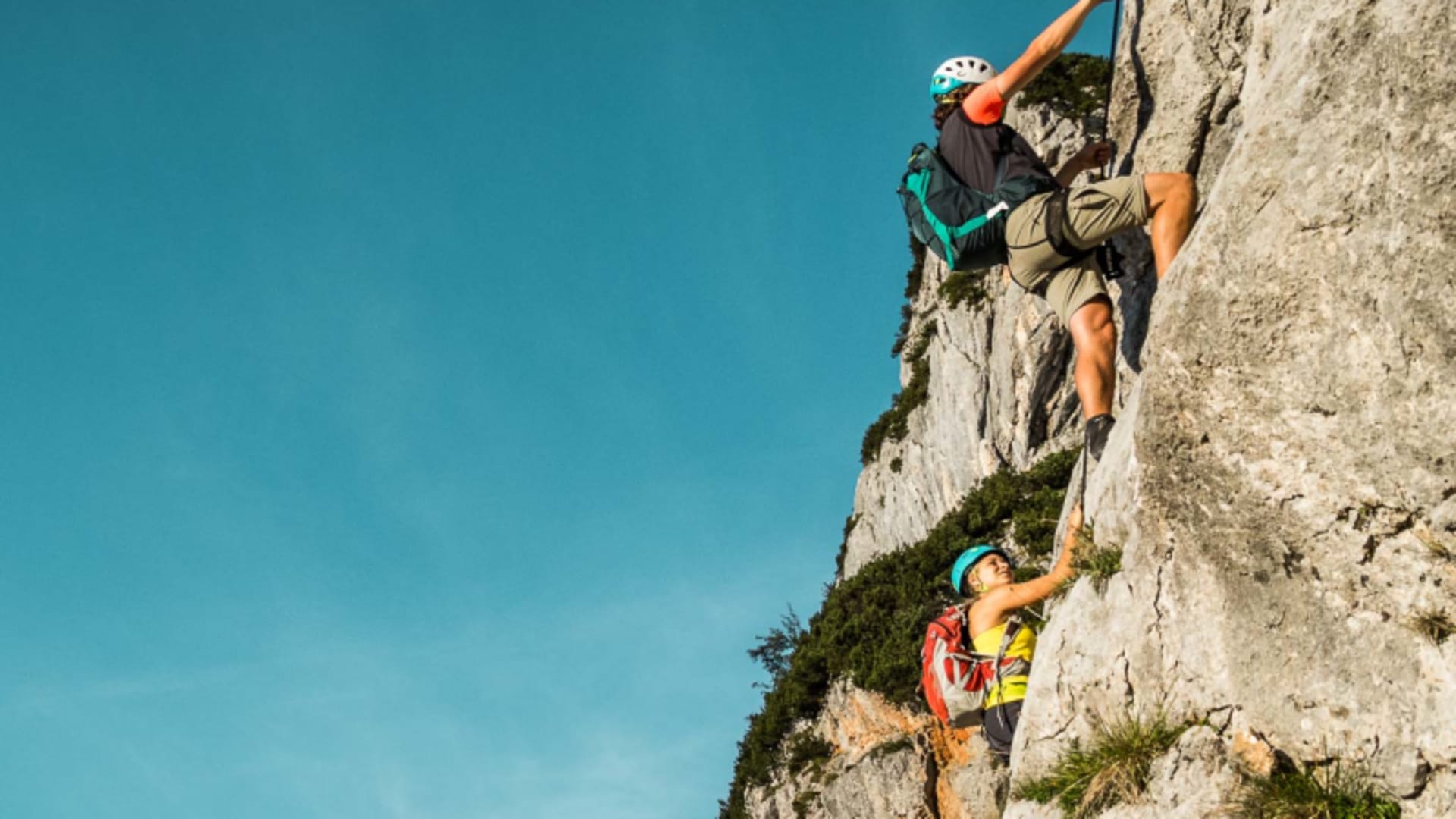 Zwei Klettersteiggeher mit Helmen steigen an einer nahezu senkrechten Kalksteinwand des Hochtronsteigs am Untersberg auf.