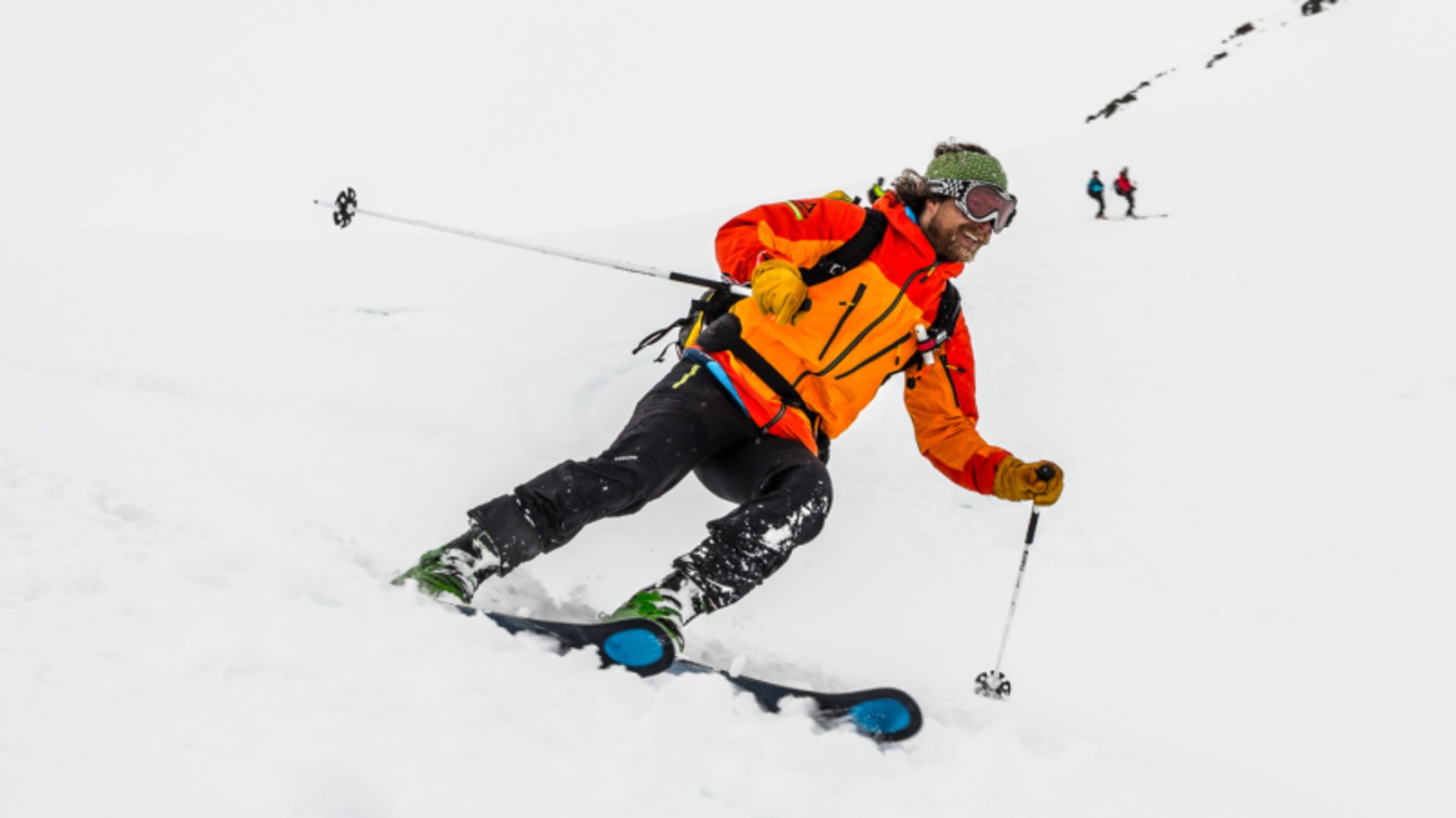 Person in orange-schwarzer Jacke mit Airbag-Rucksack fährt auf einer Skitour einen schneebedeckten Hang in den Lyngen Alps