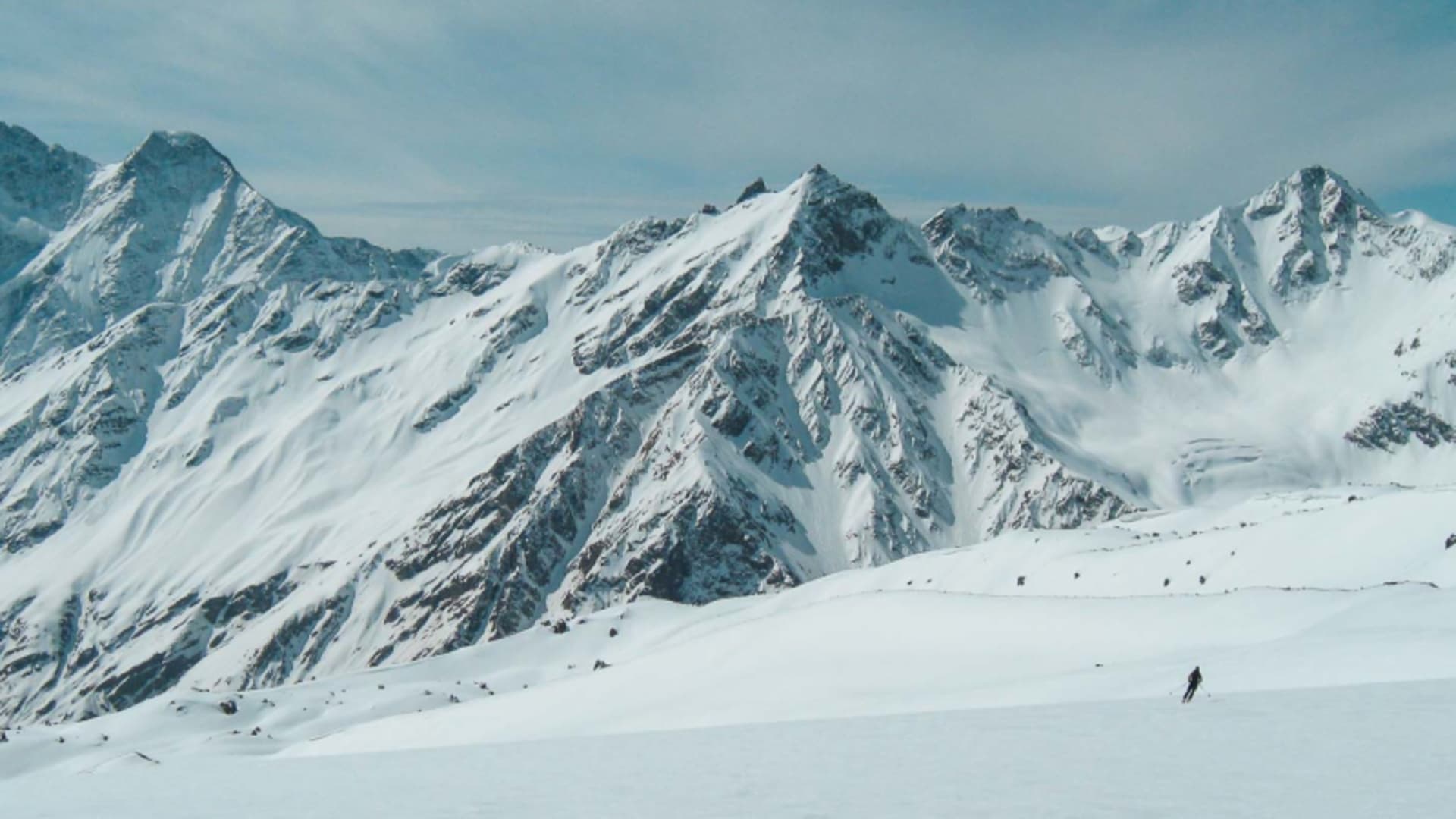 Ein Skitourengeher steigt am Elbrus im Kaukasus einen breiten Schneehang auf, im Hintergrund Fels- und Firnkämme.