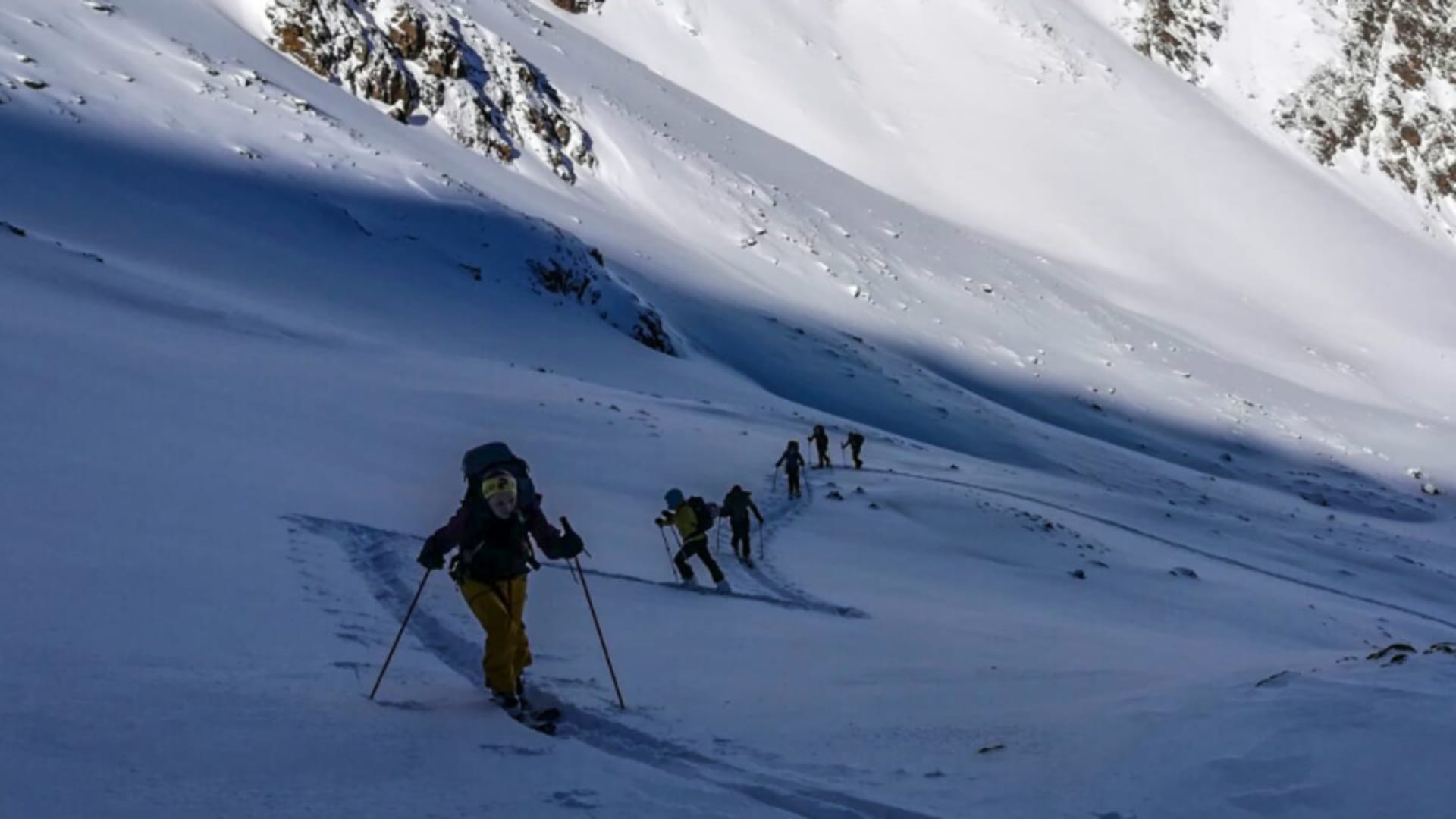 Sechs Skitourengeher steigen in Einzelreihe mit Fellen einen verschneiten Hang im Vinschgau auf.