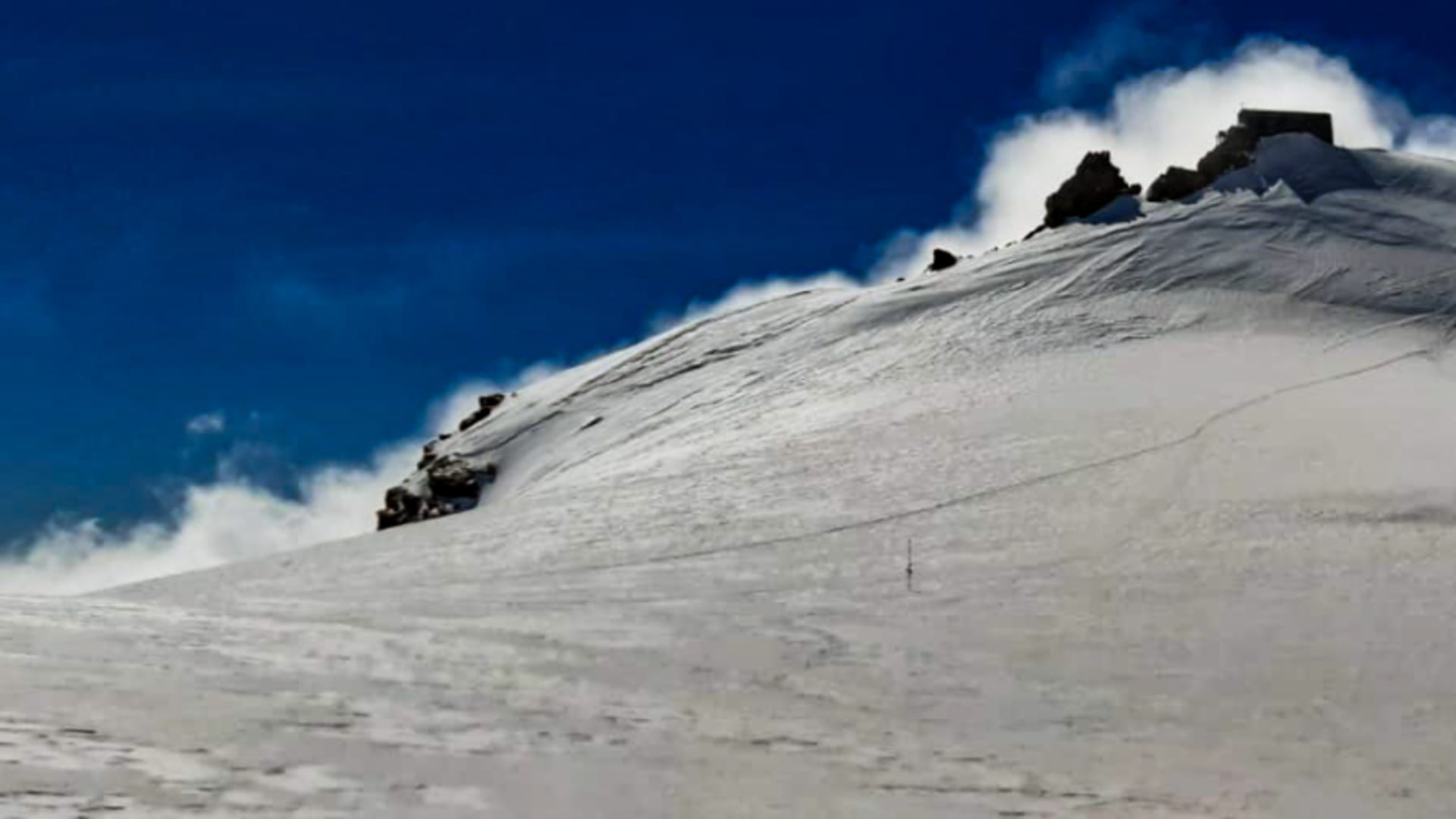 Skitour zur Capanna Margherita: schneebedeckter Steilhang mit Berghütte am Gipfel und dunkelblauen Himmel.