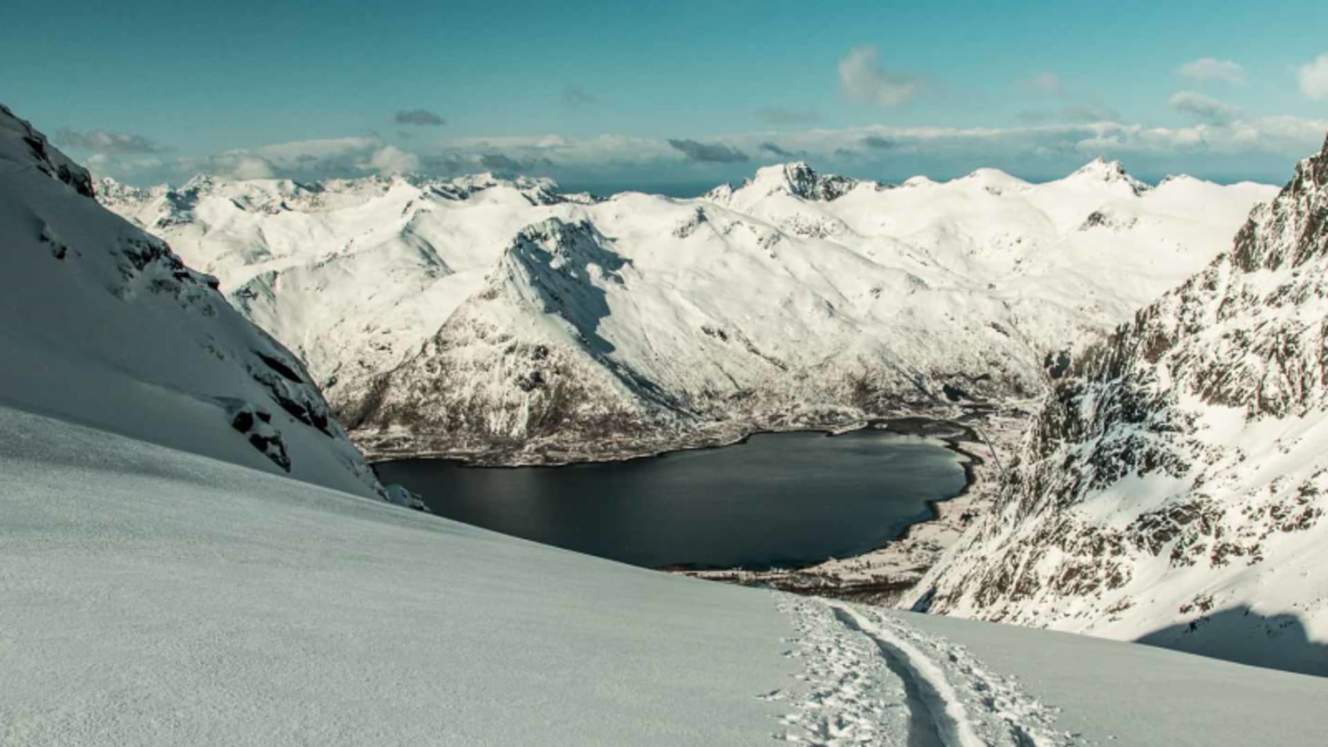 Skitour auf den Lofoten: Fellspuren auf verschneitem Hang mit Fjord und Bergkette im Hintergrund.