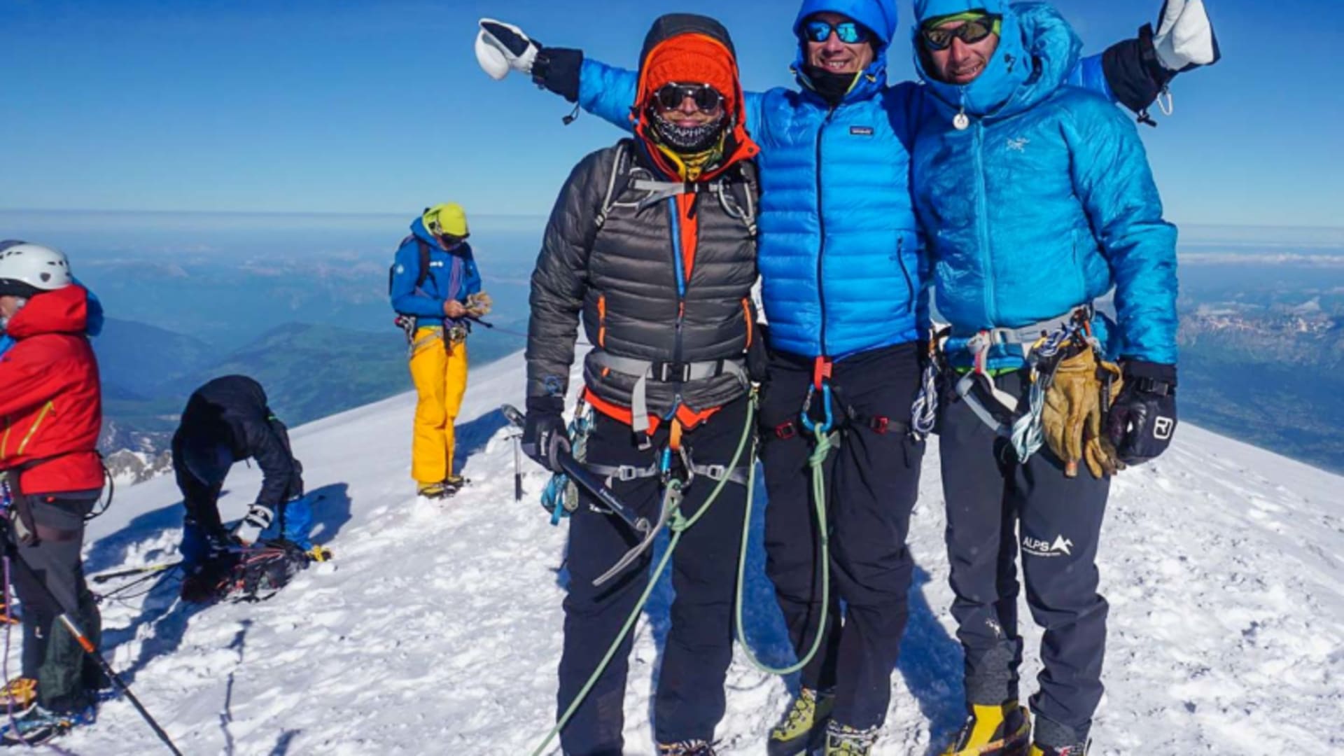 Drei Bergsteiger mit Steigeisen und Klettergurten posieren auf einem schneebedeckten Gipfelgrat im Mont-Blanc-Massiv.