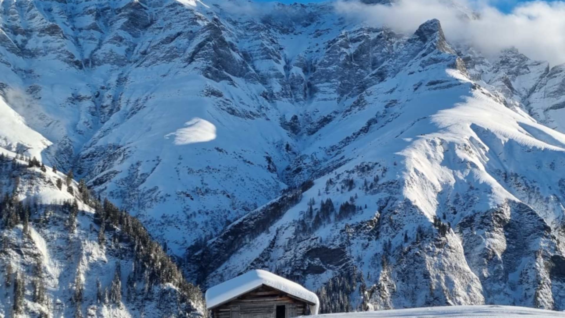 Skitour im Safiental: kleine Holzhütte mit dickem Schneedach auf weiter Schneefläche, dahinter verschneite Felsgrate