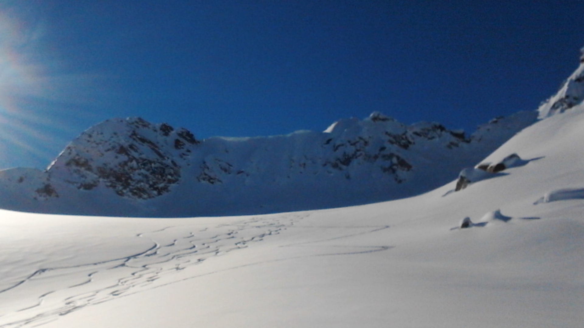 Skitour in der Silvretta: tief verschneite Hangfläche mit Spurrillen, zwei markante Gipfel und wolkenloser Himmel im Gegenlicht.