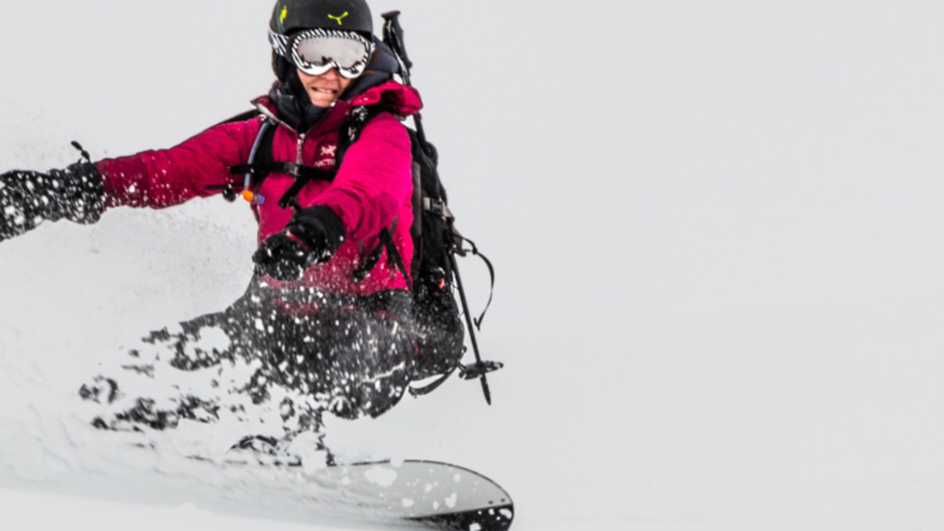 Person auf dem Snowboard in Kurvenfahrt durch Pulverschnee bei einer Bergtour in den Lyngenalpen, mit Rucksack und Skistöcken.