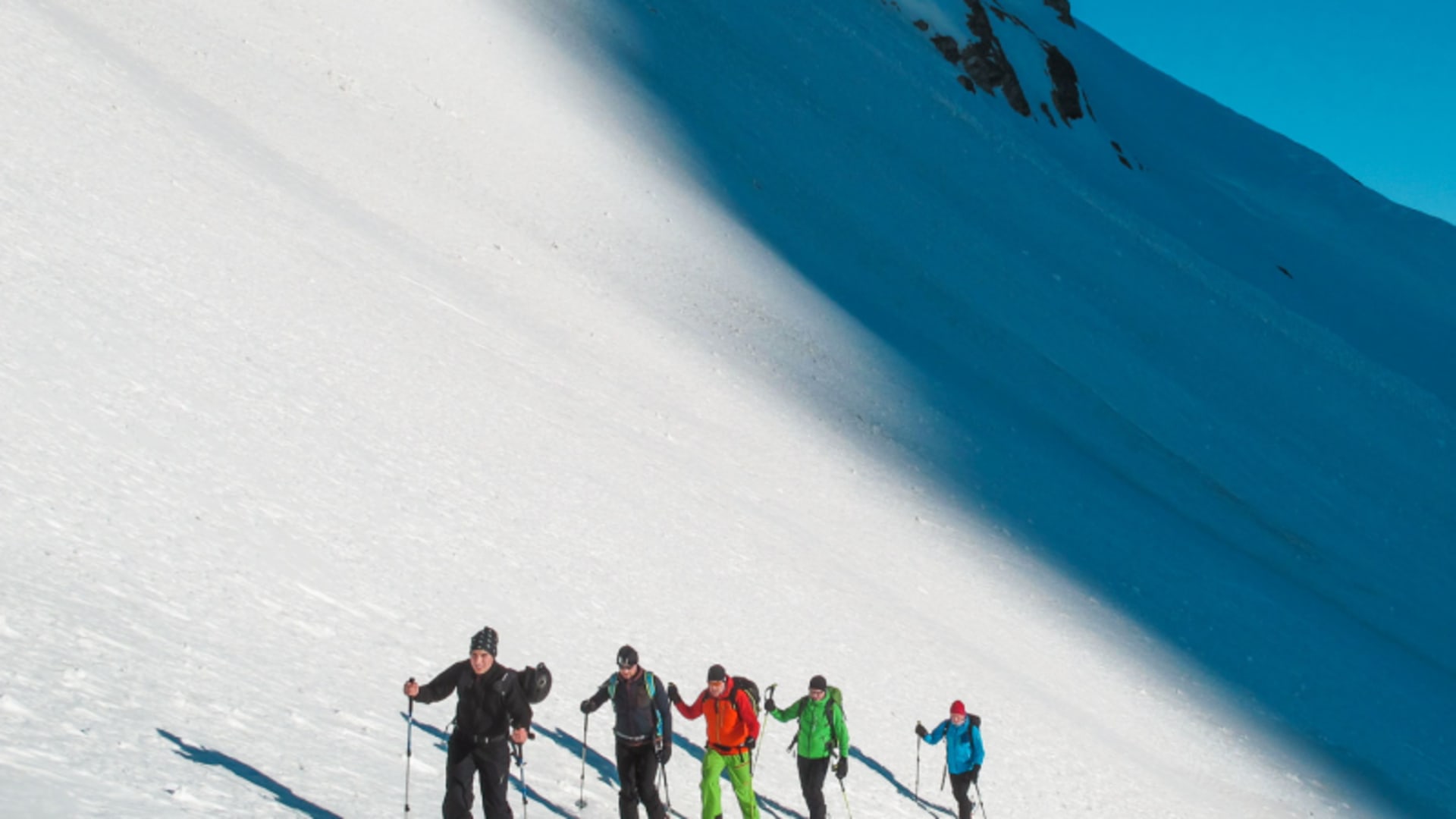 Fünf Skitourengeher üben Spuranlage in einer Reihe auf einem sonnenbeschienenen, verschneiten Berghang mit Fellen.