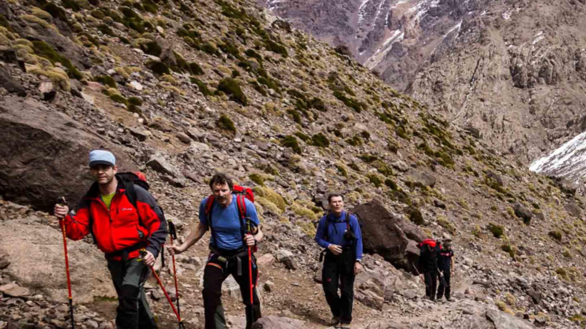 Vier Wanderer steigen mit Trekkingstöcken auf einem steinigen Pfad zur Toubkal-Hütte im Hohen Atlas auf.