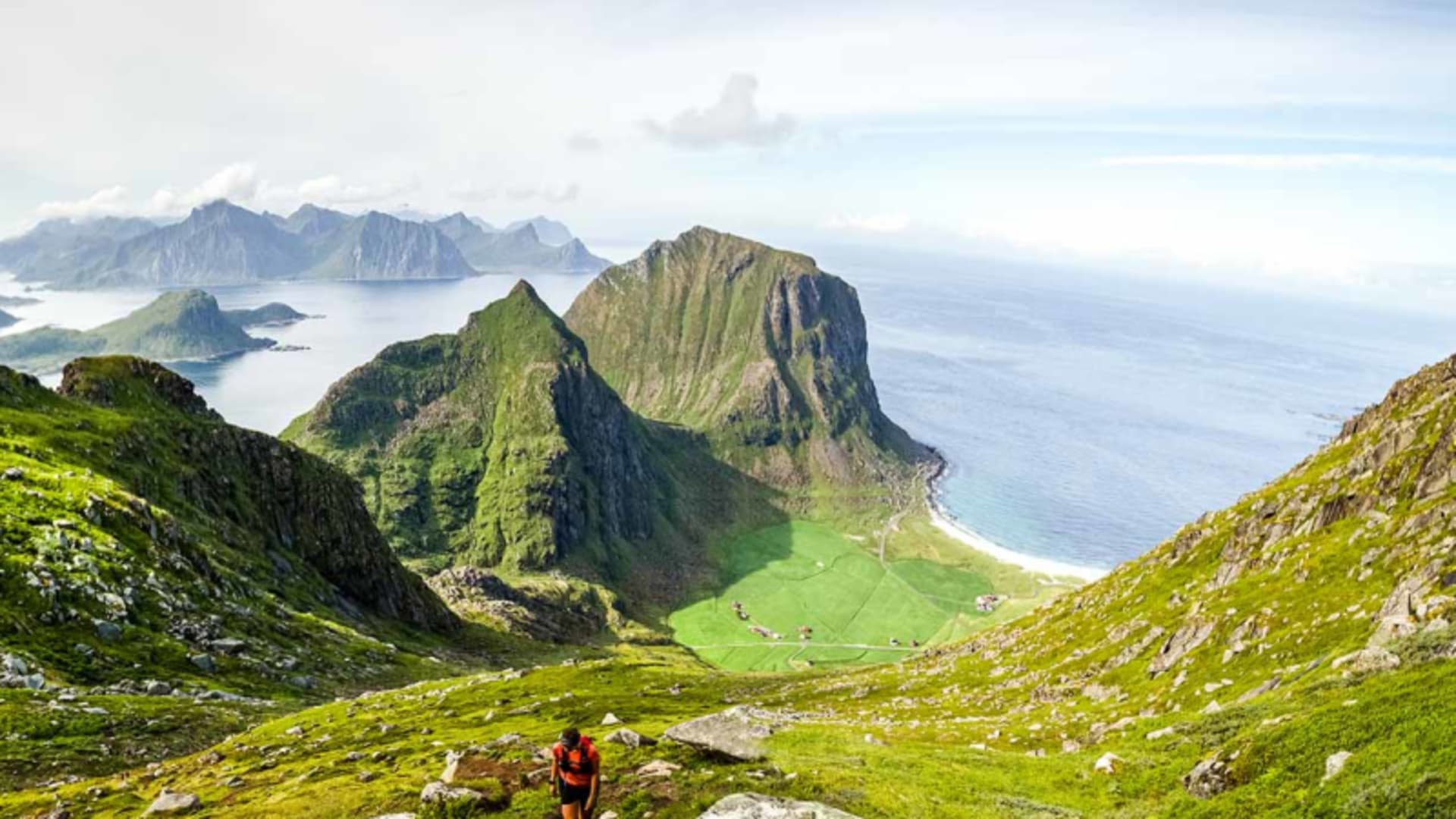 Trekking auf den Lofoten: ein Trekker mit rotem Rucksack steigt auf einem grasbewachsenen Hang mit Blick auf Meer und Inseln auf.
