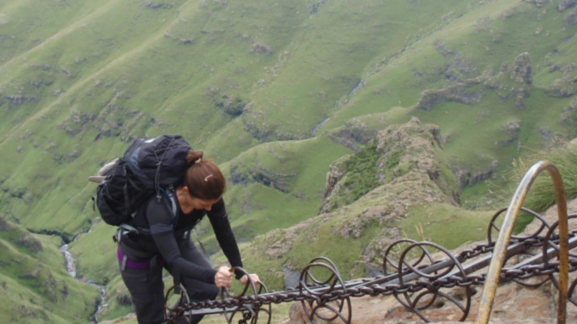 Trekking in den Drakensbergen Südafrika: Person klettert mit Rucksack an verankerten Eisenketten eine steile Felswand hinauf