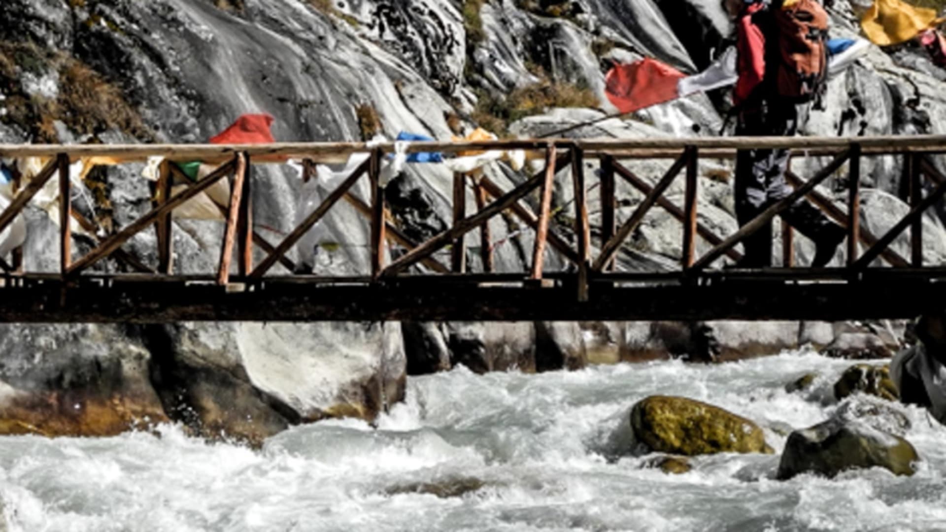 Trekking nach Namche Bazar: ein Trekker überquert eine mit Gebetsfahnen geschmückte Hängebrücke über einen Gebirgsfluss.