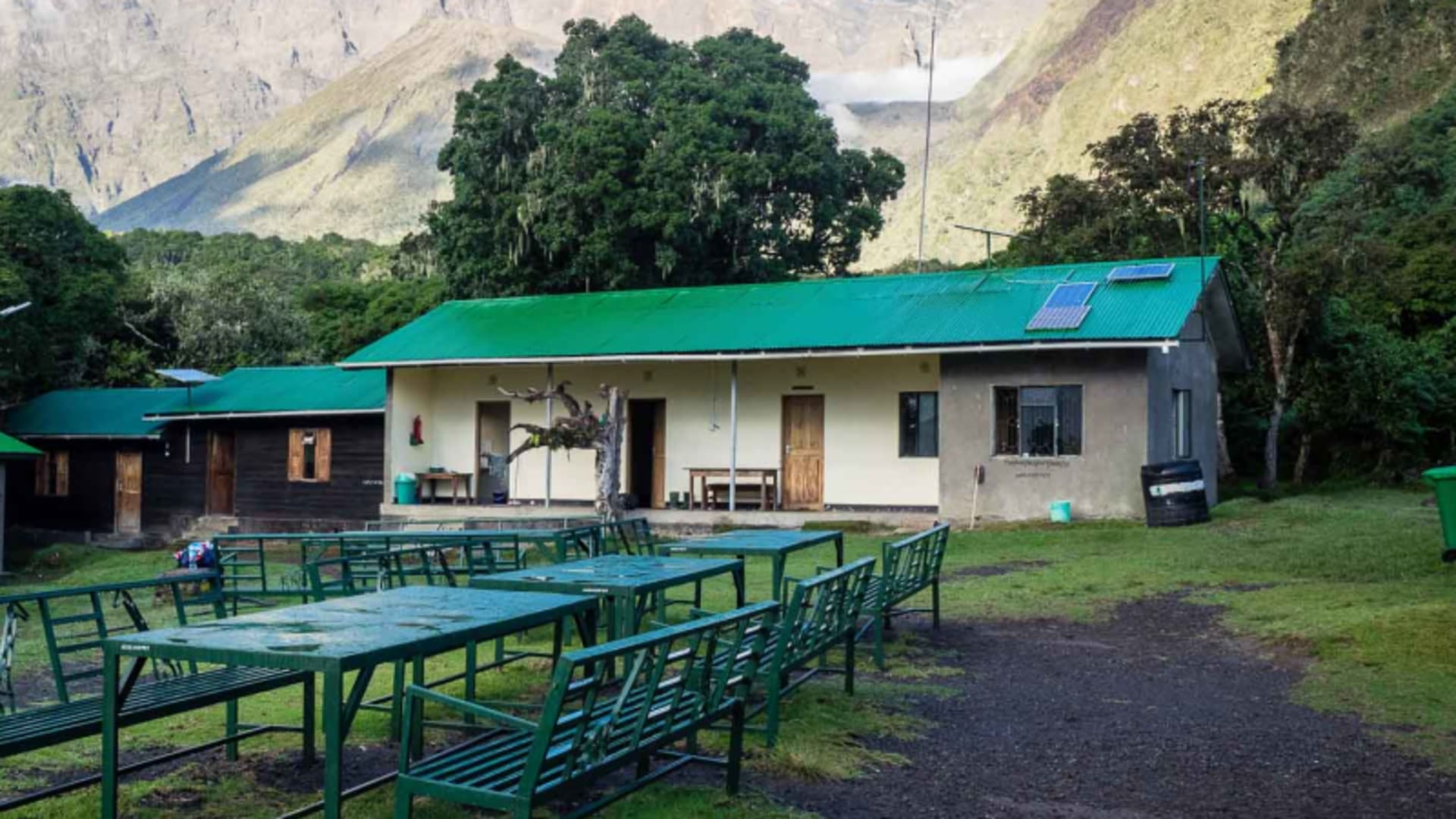 Trekking am Kilimandscharo: Hüttenkomplex mit Metalltischen und Solarpanelen vor steiler Felsbergkette.