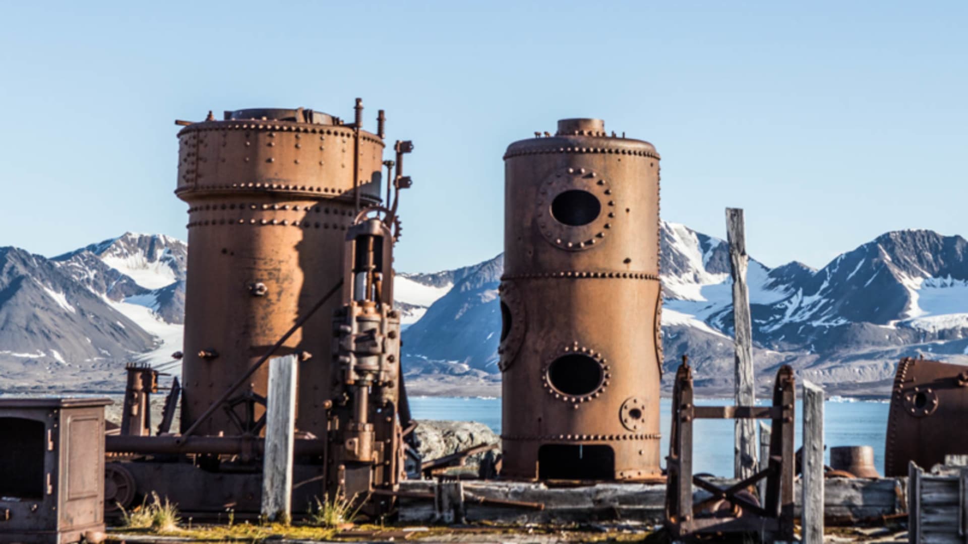 Trekking auf Spitzbergen: verrostete Industriekessel und Maschinenteile einer verlassenen Anlage vor Fjord und Bergpanorama.
