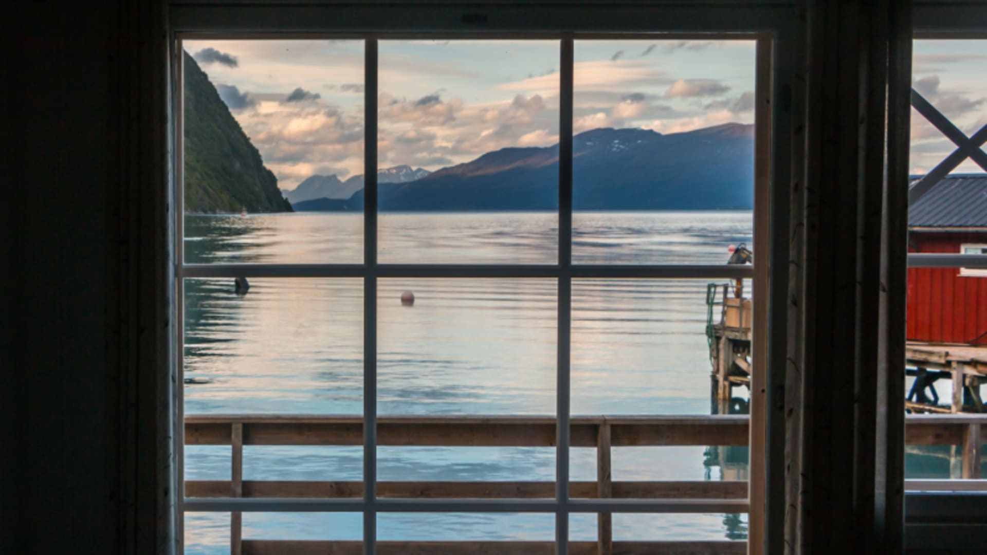 Blick durch ein Sprossenfenster einer Trekking-Lodge in den Lyngenalpen auf einen spiegelglatten Fjord mit schneebedeckten Bergen