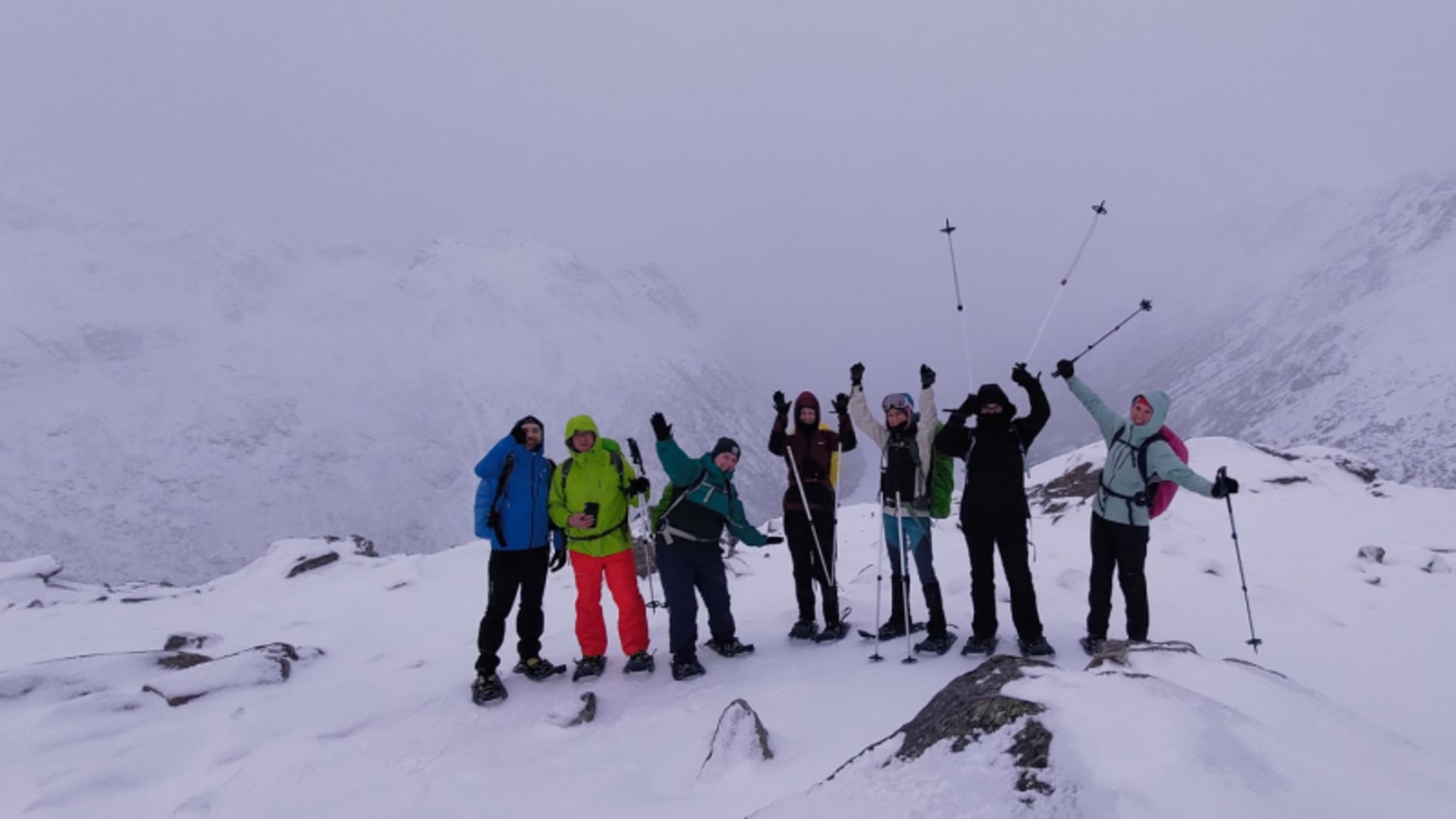 Acht Winterwanderer jubeln mit erhobenen Armen und Stöcken auf einem schneebedeckten Lofoten-Gipfel.