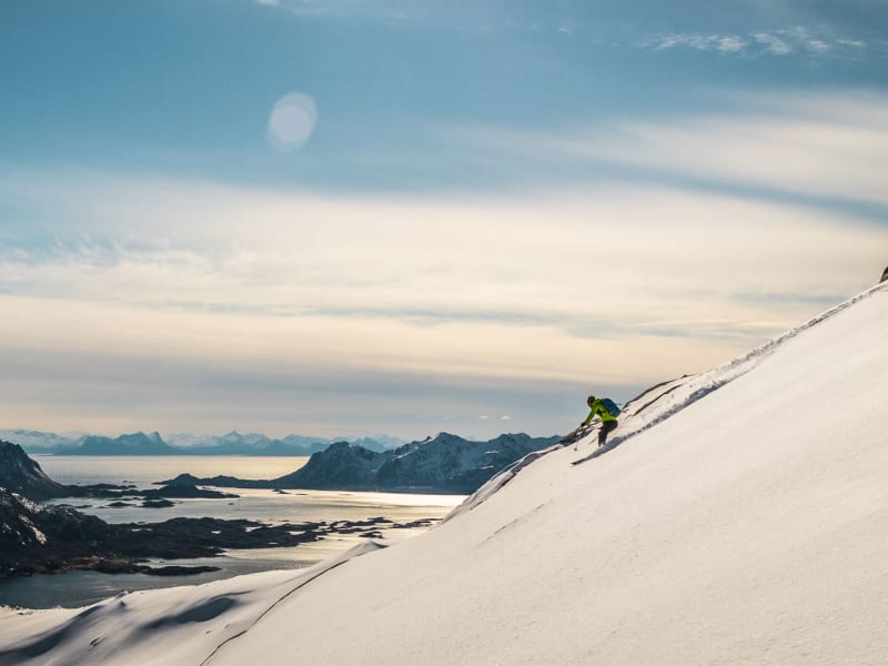 Skitourengeher in Abfahrtshaltung auf steilem Hang mit Fjord und Schären im Hintergrund auf den Lofoten.