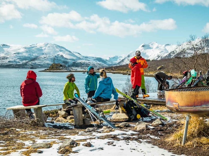 Skitourengehergruppe pausiert auf einer Bank am Fjordufer der Lofoten mit Ski und Bergen im Hintergrund.