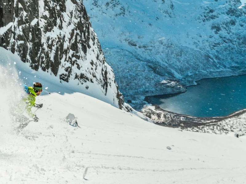 Skitourengeher fährt in gelbgrüner Jacke einen steilen Pulverschneehang mit Fjordblick auf den Lofoten ab.