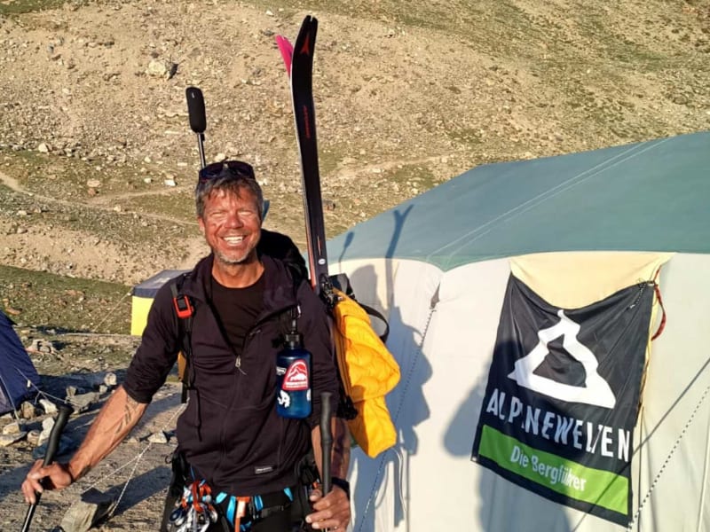 Freudestrahlender Gipfelbesteiger mit Rucksack und Skistöcken vor Zeltlager auf steinigem Gelände bei einer Bergtour.