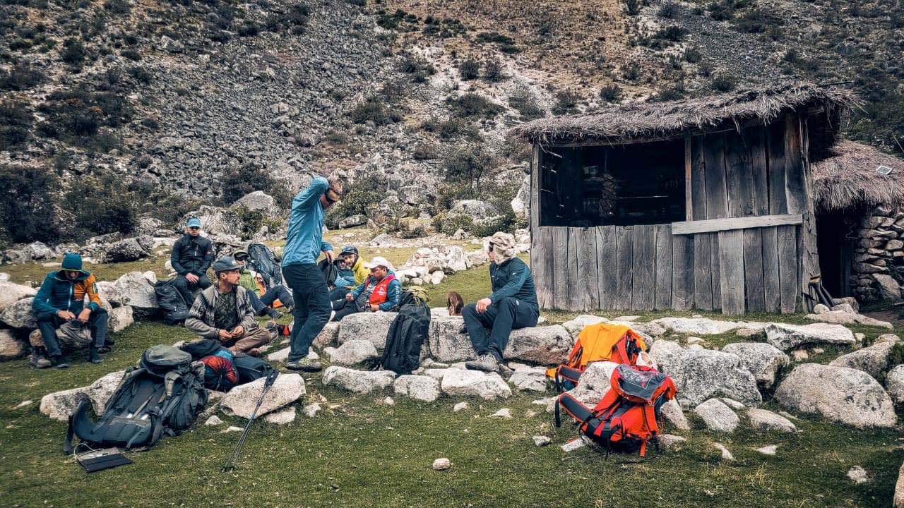 Bergtour am Alpamayo: Gruppe von Bergsteigern rastet auf Felsen neben einer Holzhütte mit Grasdach.