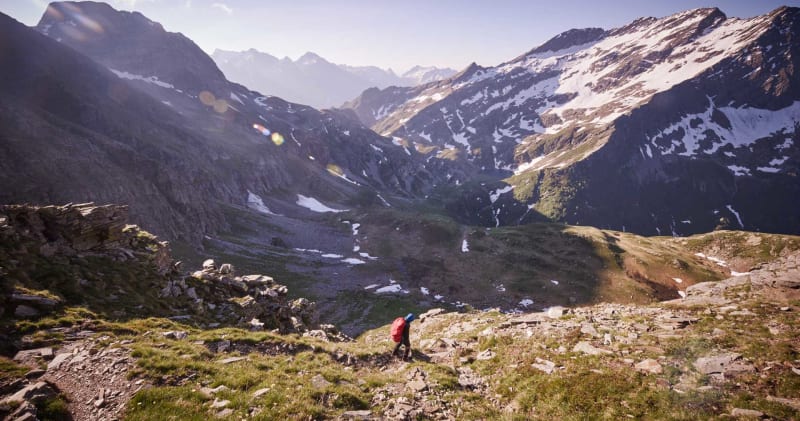 Bergtour allein auf weiter Flur: Bergsteiger mit rotem Rucksack steigt einen felsig-grasigen Hang in einem alpinen Hochtal