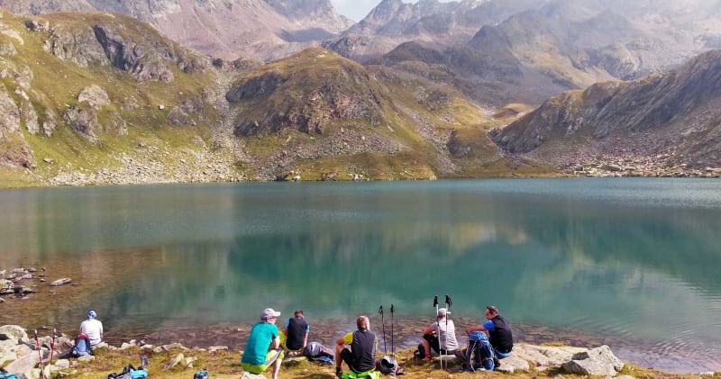Sieben Bergsteiger rasten am türkisgrünen Bergsee bei Meran mit Rucksäcken auf Felsen.
