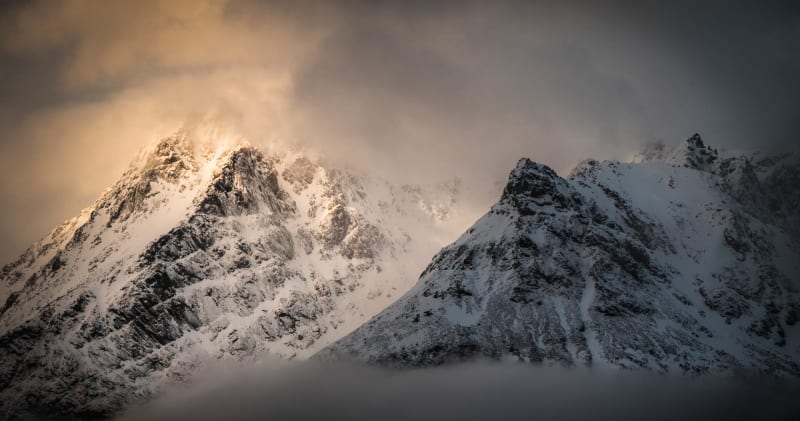 Schneeschuhtour auf den Lofoten: zwei stark verschneite Berggipfel im Wolkenband mit warmem Lichteinfall