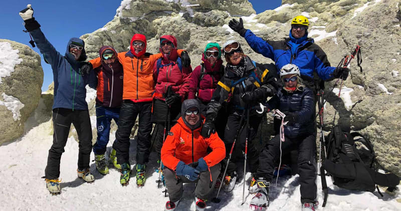 Zehn Skitourengeher posieren mit Steigeisen und Stöcken am felsigen Gipfel des Damavand im Iran.