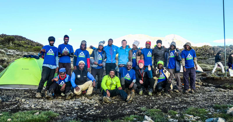Trekking am Kilimandscharo: Gruppe von ca. 18 Trekkern im Hochlager vor schneebedecktem Gipfel.