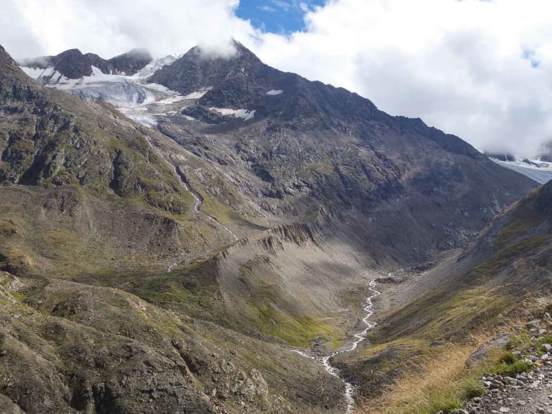 Bergwanderung in der Bernina: Schotterweg durch karges Hochtal mit mäanderndem Gletscherbach und Gletscherzungen im Hintergrund.