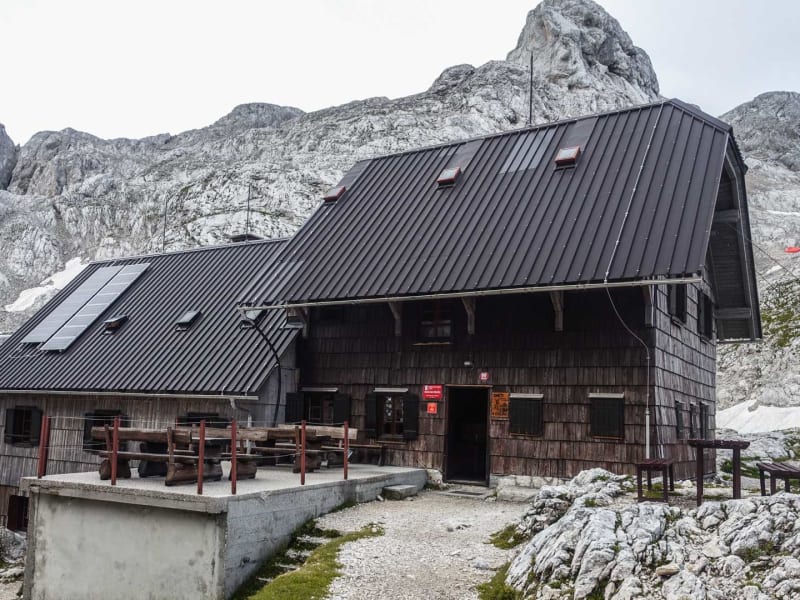 Bergwanderung in Slowenien: Holzberghütte Koča na Dolicu vor verkarsteten Kalkfelsen mit Schneeresten.