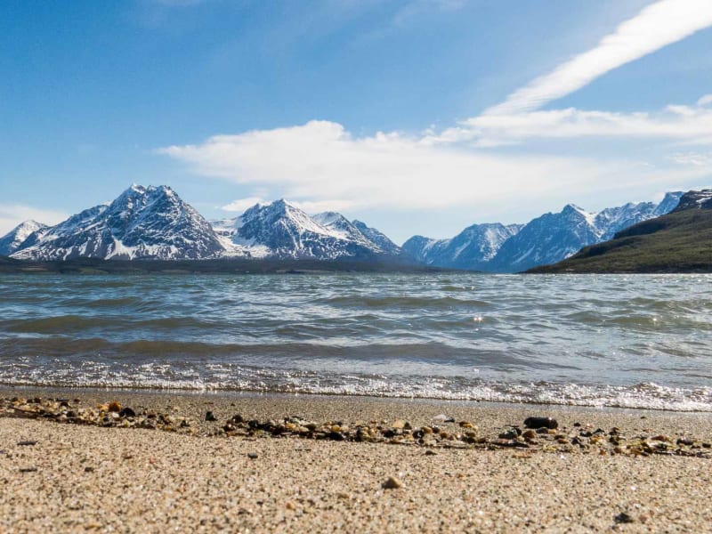 Bergwanderung Lyngenalpen Norwegen: Sandstrand mit Brandungswelle, blaugrünem Fjordwasser und schneebedeckten felsigen Gipfeln