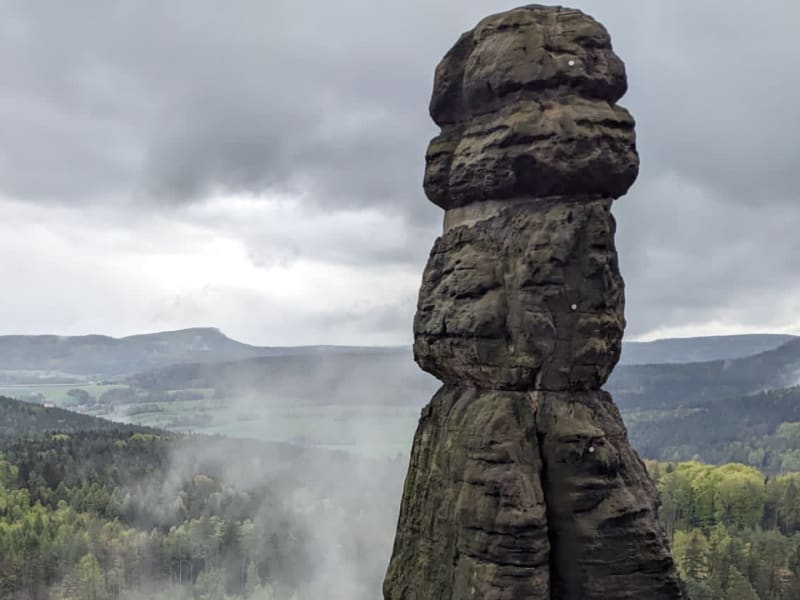 Bergwanderung in der Sächsischen Schweiz: schlanker Sandsteinfelsen mit Kletterbolzen vor bewaldeter Tallandschaft.