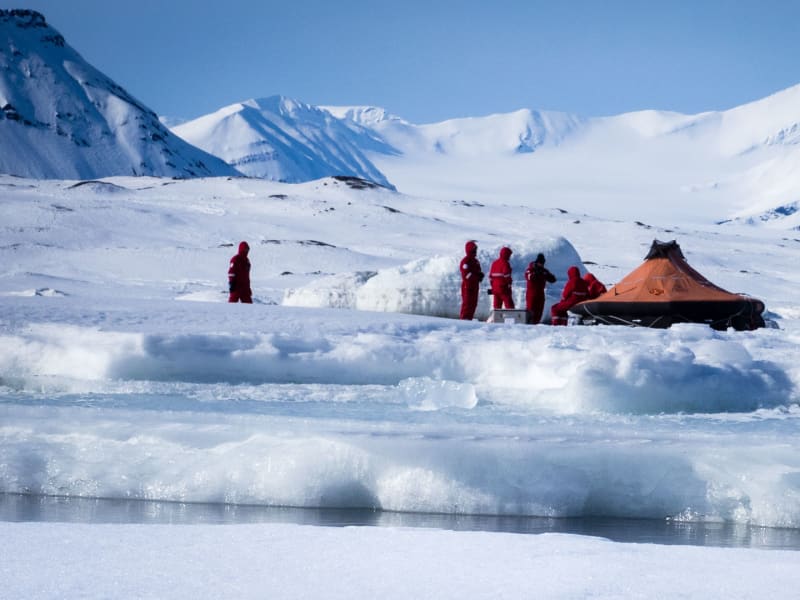 Expedition auf Spitzbergen: fünf Höhenbergsteiger in roten Schutzanzügen neben Rettungsinsel und Schneebau auf Packeis.