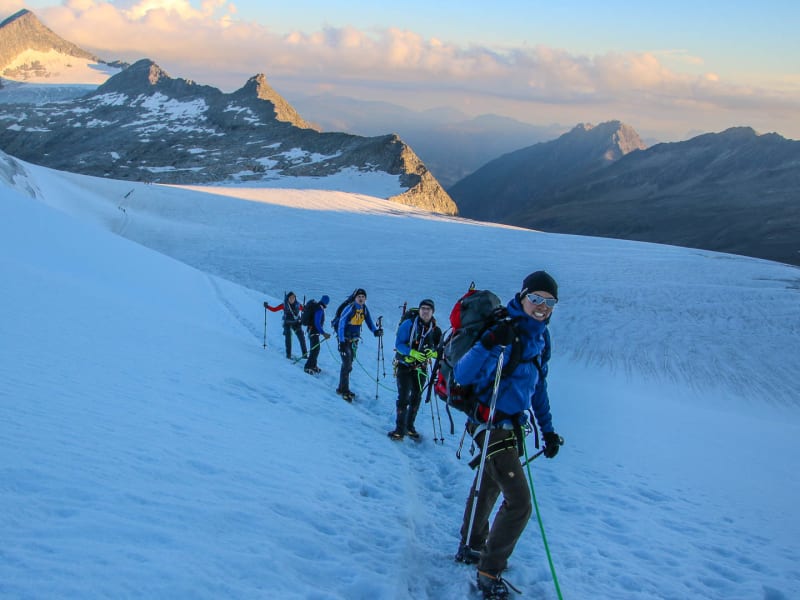 Fünf Bergsteiger mit Steigeisen und Rucksäcken steigen im Morgen- oder Abendlicht einen Schneehang zum Großvenediger auf.