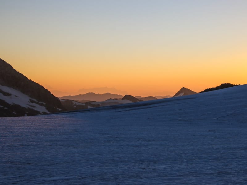Schneebedeckter Hang am Großvenediger bei Sonnenaufgang mit orangegelbem Lichtschein und Bergsilhouetten im Hintergrund.