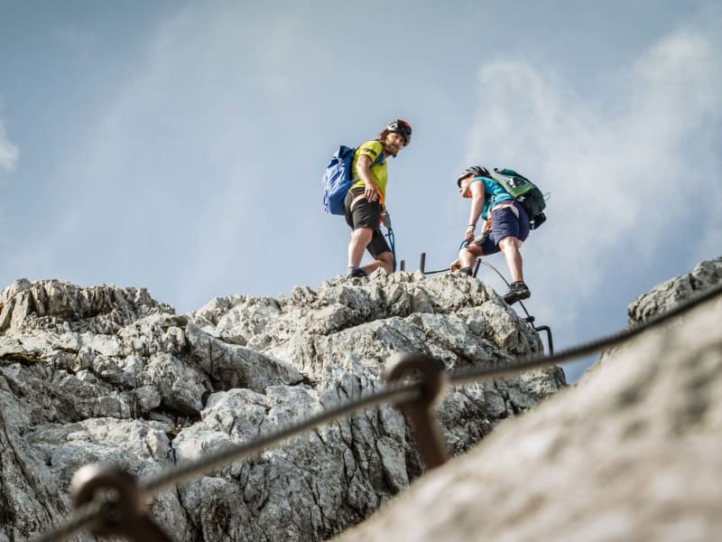 Zwei Klettersteiggeher steigen am Kalkfels der Alpspitze aufwärts, Stahlseil und Eisenstifte im Vordergrund