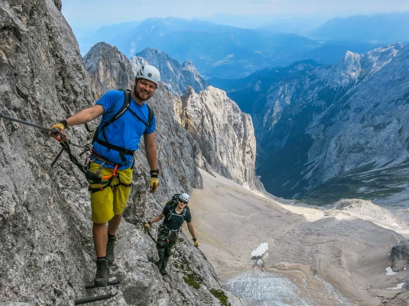 Zwei Klettersteiggeher an Eisenbügeln im Höllental, im Hintergrund der Gletscherrest und das Zugspitzmassiv.