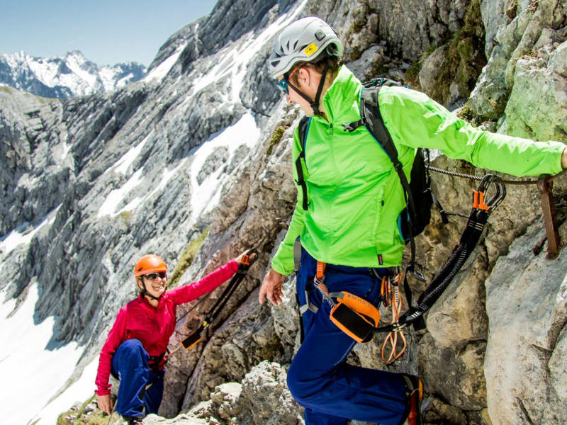 Zwei Klettersteiggeher mit Klettergurt und Karabinern greifen am Drahtseil einer Felswand beim Kreuzeckhaus
