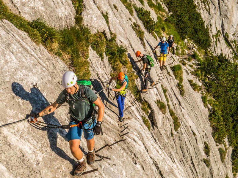 Fünf Klettersteiggeher traversieren an Eisenstiften und Fixseil eine senkrechte Kalkfelswand am Klettersteig zur Zugspitze.