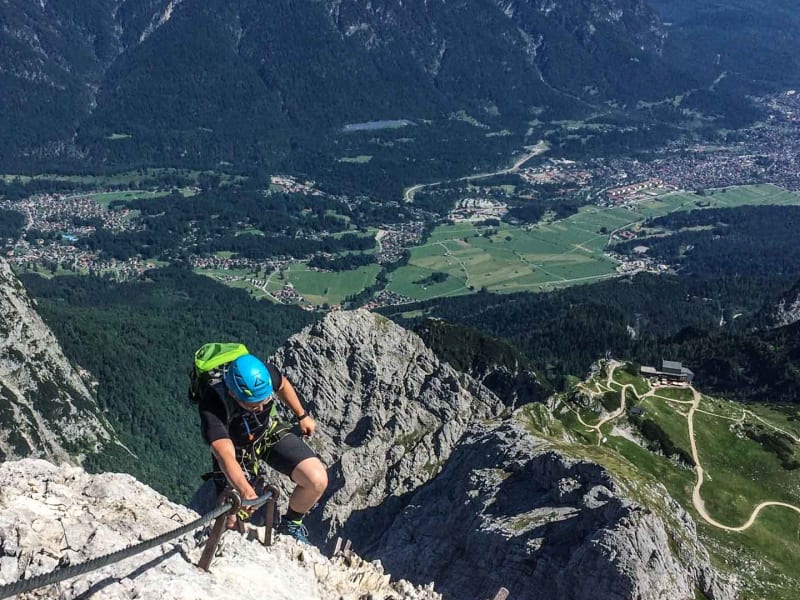 Klettersteig an der Alpspitze: Klettersteiggeher greift an Eisenklammern einer Kalkfelswand, Tal im Hintergrund.