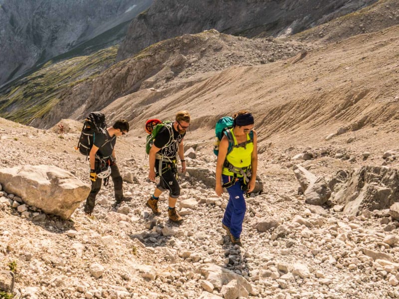 Drei Klettersteiggeher mit Gurten und Helmen steigen über steiles Geröll zwischen dem Brett und dem Höllental-Gletscher