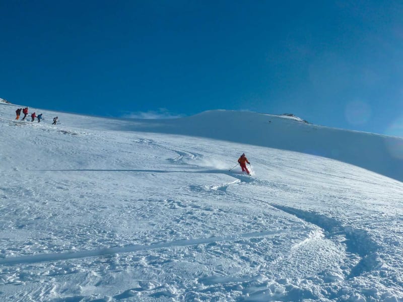 Skitourengeher in roter Jacke fährt Tiefschnee ab, vier weitere Skitourengeher steigen an der Heidelberger Hütte auf.