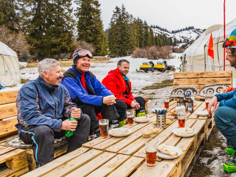 Vier Skitourengeher rasten an einem Holzpalettentisch vor einer weißen Jurte im verschneiten Winterlager in Kirgistan.