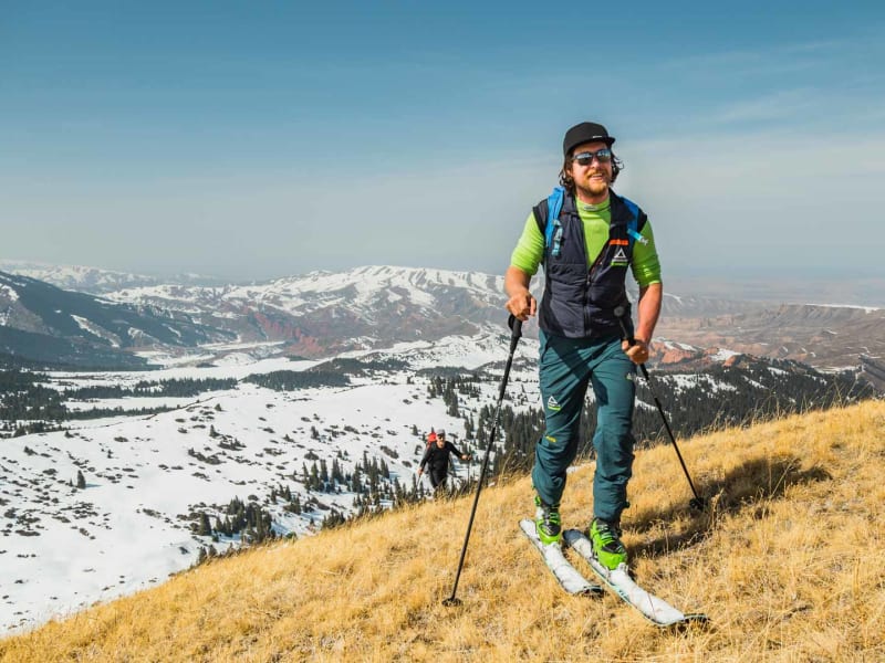 Drei Skitourengeher im Aufstieg auf trockenem Grashang in Kirgistan, schneebedeckte Bergrücken im Hintergrund.