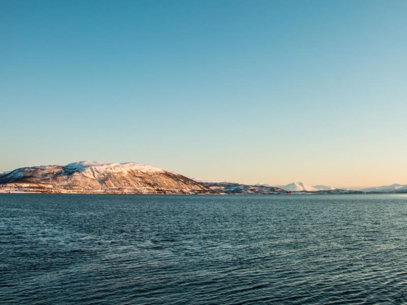 Skitour per Segelschiff in Norwegen: winterliches Fjordpanorama mit schneebedeckten Bergen im warmen Morgen- oder Abendlicht.