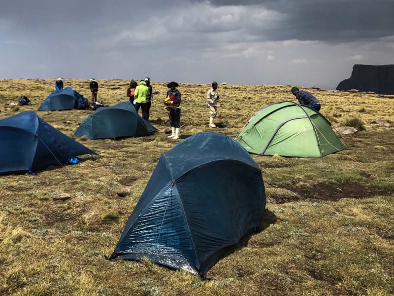 Trekking in den Drakensbergen: Zeltcamp auf dem Hochplateau mit vier Kuppelzelten und Trekkergruppe bei Gewitterwolken.